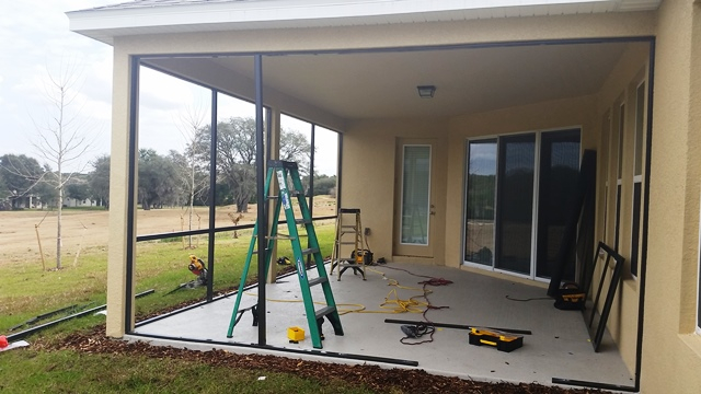 A partially constructed screened-in patio with tools, a green ladder, and wiring on the concrete floor. The patio is attached to a beige house with large windows and overlooks a grassy yard with trees.