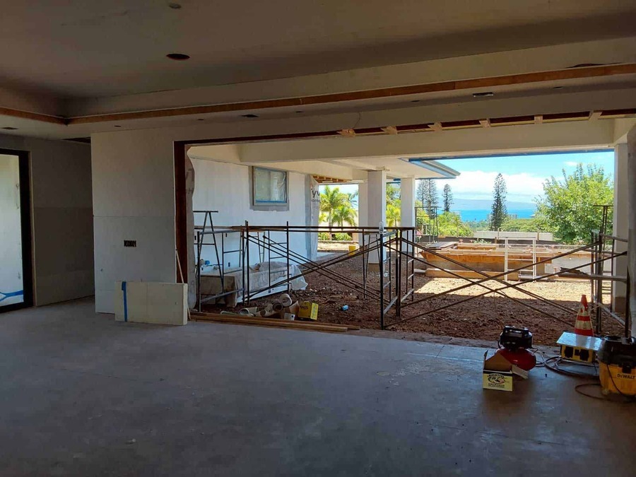 View from inside a partially constructed building with an open wall, scaffolding, and construction materials. Outside, there are trees, plants, and a distant view of the sea under a blue sky.