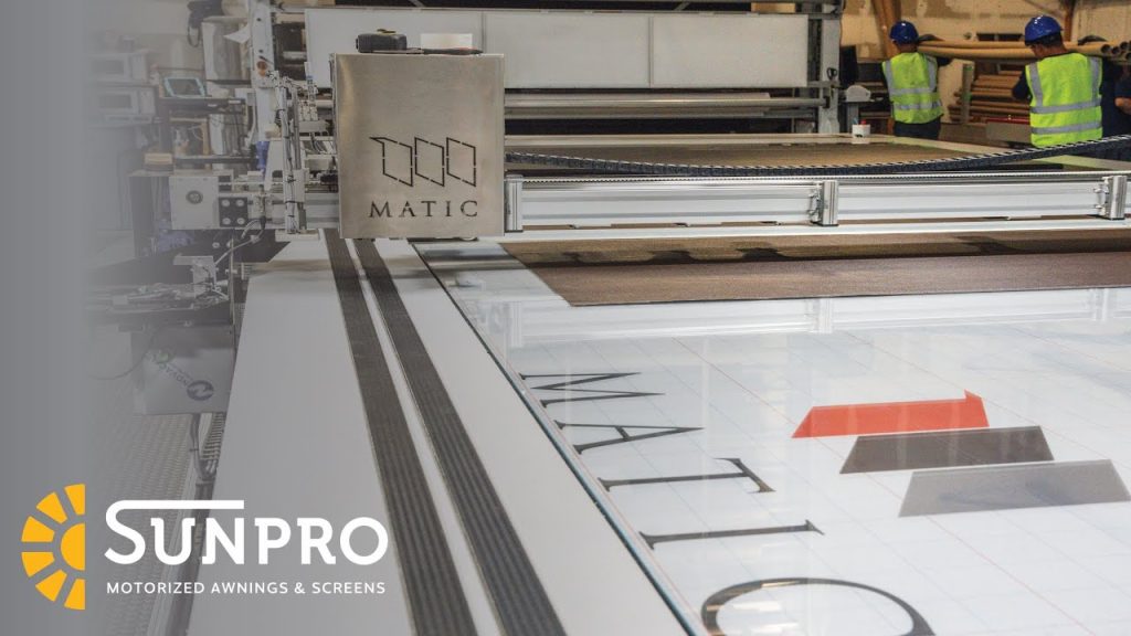 A large industrial printer prints the word “MATIC” and colored shapes on a wide sheet. Two workers in safety vests and helmets stand in the background. The SunPro Motorized Awnings & Screens logo is in the lower left corner.