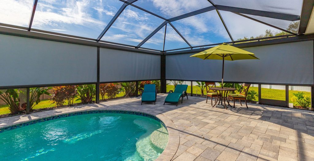 Sunpro motorized Screened-in outdoor pool area with two blue lounge chairs, a round table with four chairs, and a green umbrella on a stone patio. Sunlight shines through the screened roof and side panels.