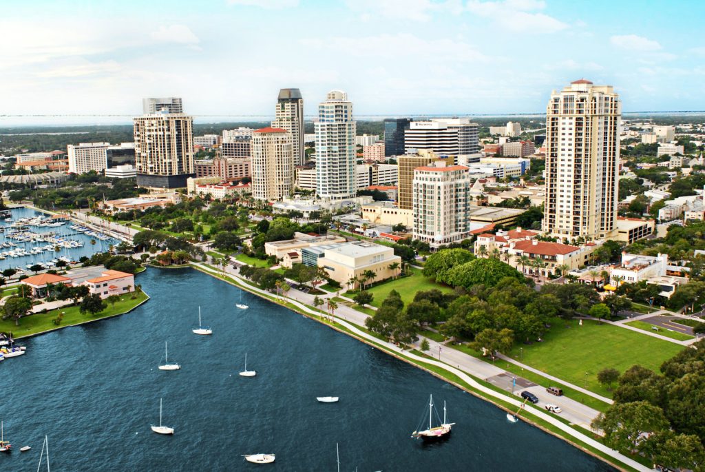 Aerial view of a coastal city with tall buildings, tree-lined streets, and a waterfront park featuring retractable awnings. Boats are anchored in a marina beside blue water under a partly cloudy sky—scenes typical of Fort Myers, FL.