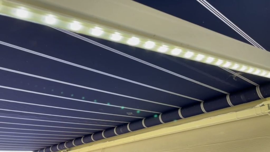 A close-up view of a blue retractable awning with white stripes, seen from below, with a strip of bright LED lights attached along the edge.