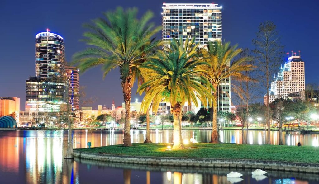 Night view of downtown buildings illuminated behind palm trees and a small grassy island in a lake, with reflections on the water and swans swimming in the foreground. The vibrant cityscape showcases outdoor spaces enhanced by Central Florida Awnings, adding charm and functionality to the waterfront ambiance.