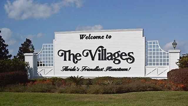 White sign with black text reads, Welcome to The Villages, Florida’s Friendliest Hometown! It is surrounded by beautiful landscaping, lanterns, and a blue sky in the background—showcasing the charm that makes this area perfect for features like Central Florida Awnings.